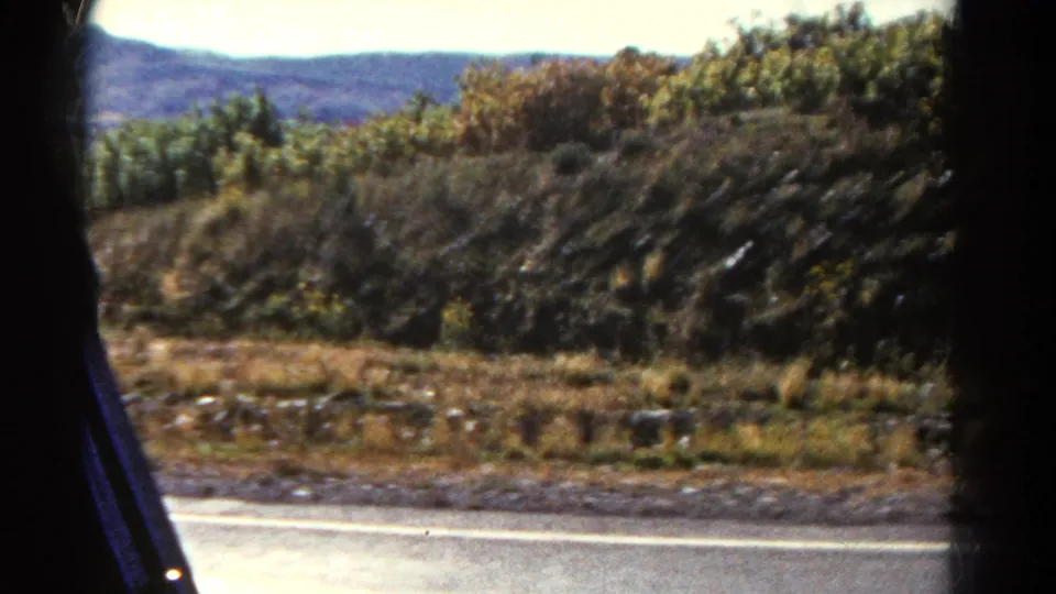 1967 Colorado car-window view with dry roadside vegetation, rocky ground, low hills, and distant ridgeline under bright sky.