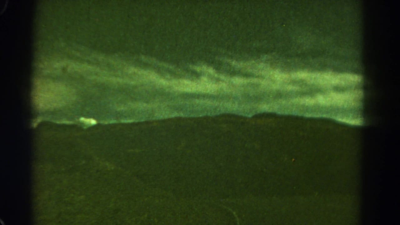 1967 8mm Colorado landscape frame showing a green cast, cloudy sky, dark distant ridge, and faint pale object near the left horizon.