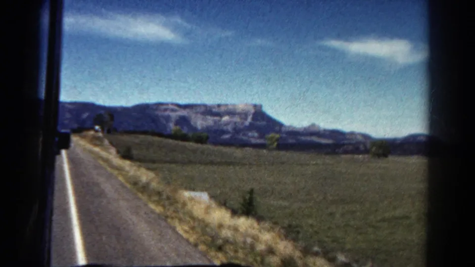 1967 Colorado road-trip view from a moving vehicle, showing a paved road, open fields, distant mesa, and blue sky.