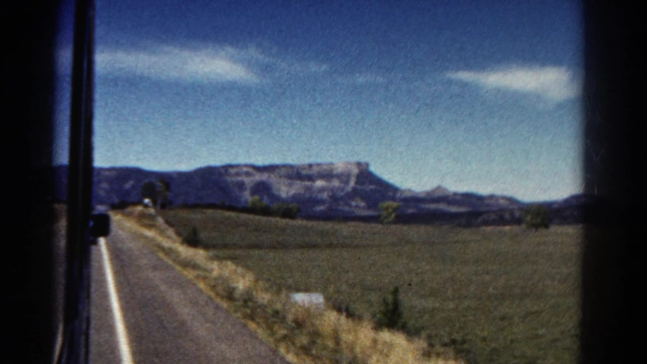 1967 Colorado 8mm frame from a moving vehicle showing a paved road, open fields, scattered trees, and distant mesa-like mountains.