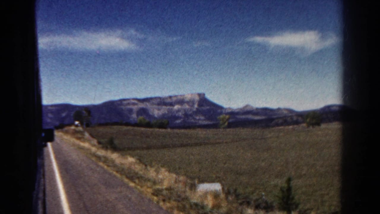 Passenger-view 8mm frame of rural Colorado countryside with a paved road, open fields, trees, and a flat mountain ridge on the horizon.