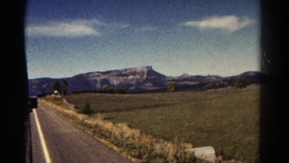 1967 Colorado home-movie frame showing a road-edge view across fields toward mesa-like mountains, with visible 8mm grain and gate edges.