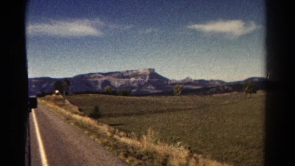 Final 8mm frame of the Colorado drive with a paved roadside, broad fields, scattered trees, and distant flat-topped mountains.