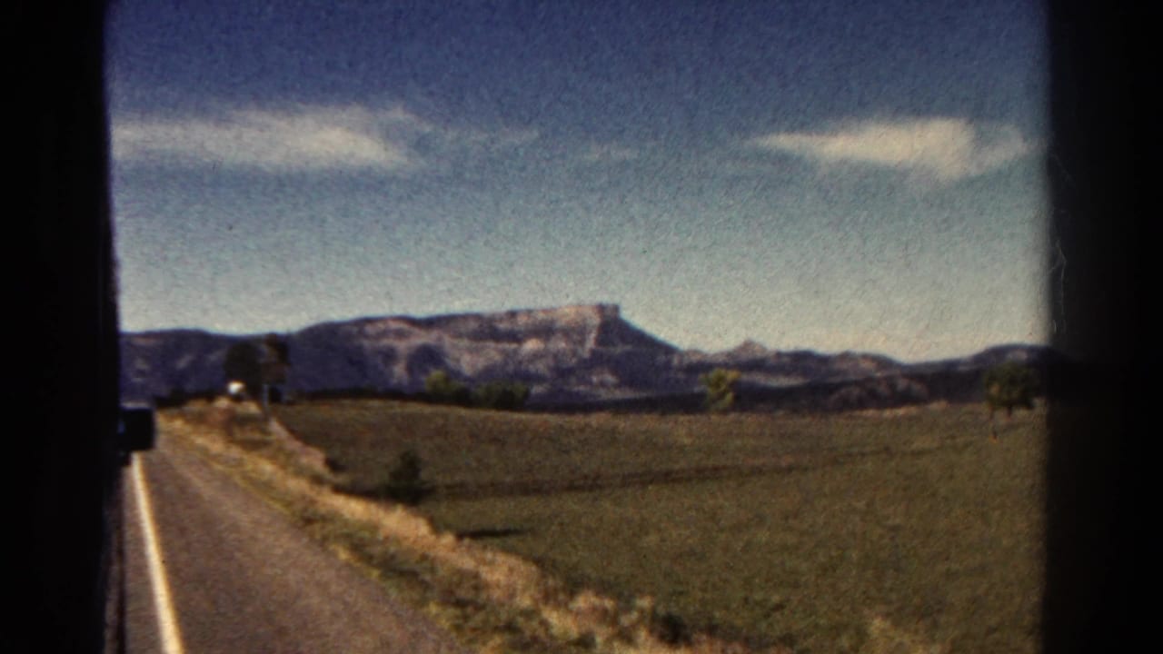 1967 Colorado 8mm frame showing a passenger-side road view with open fields, dry grass, distant ridges, and a flat-topped mountain beneath light clouds.