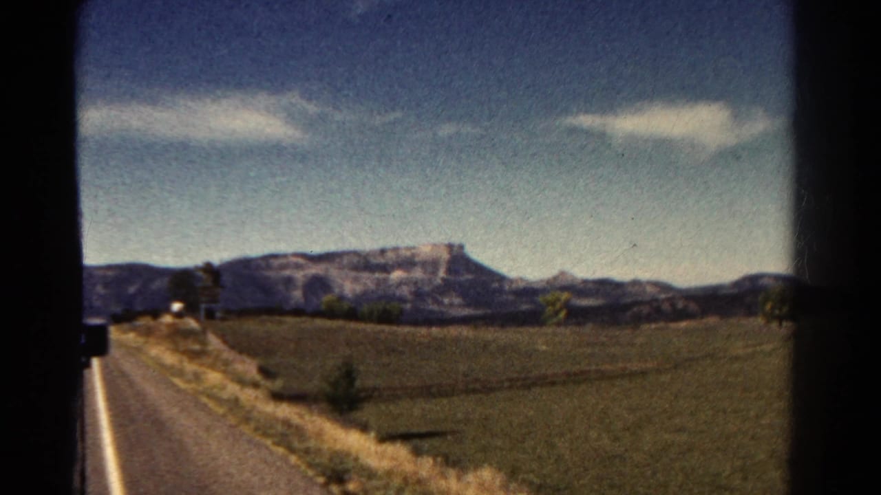 1967 Colorado road-trip frame with the road running along the left edge, open agricultural-looking land in the foreground, and low mountains in the distance.