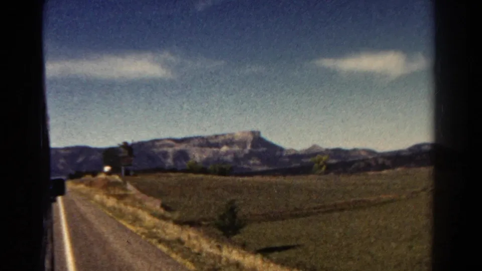 1967 Colorado 8mm still showing a rural road trip view with dry verge, broad field, scattered trees, and a distant mesa-like mountain form.