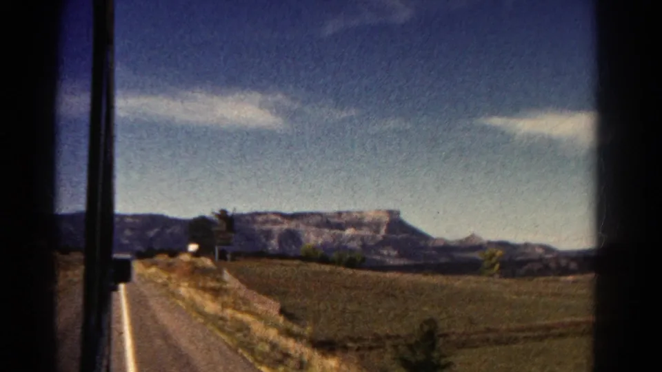 1967 Colorado home-movie frame with car-window borders framing a sunlit field, a roadside strip, trees, and a distant flat-topped mountain under a bright sky.