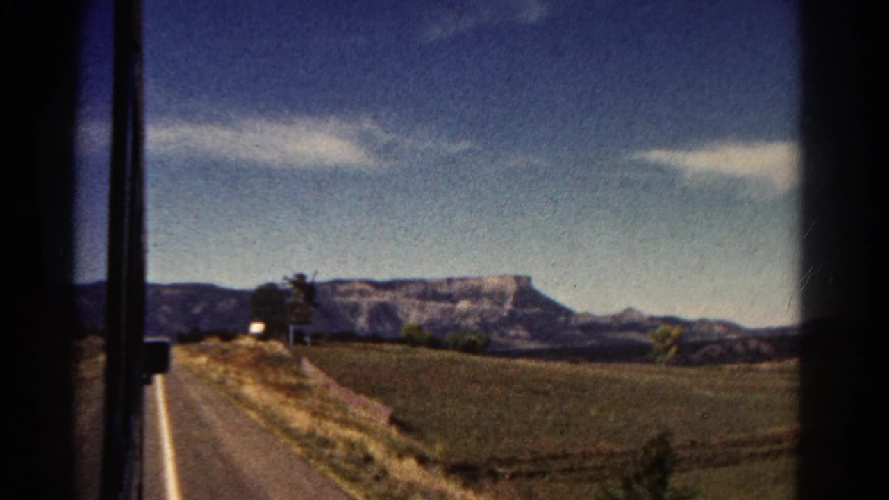 Road beside open fields with distant flat-topped mountains under a blue sky.