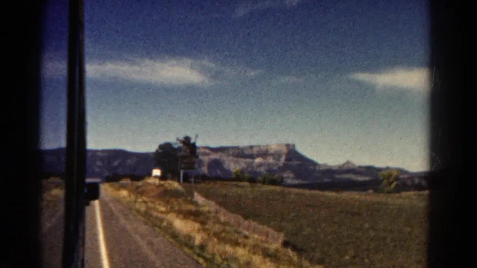 1967 Colorado home-movie frame with a road at left, open fields, roadside trees, and a distant mesa under bright sky.