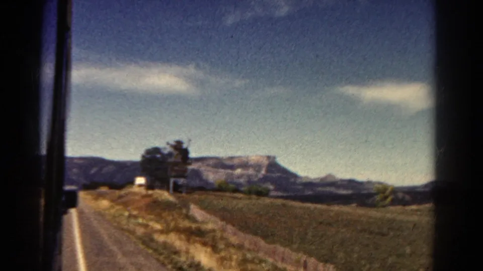 Window-framed Colorado landscape with a paved road, open field, brush, and mesa-like mountains in the distance.