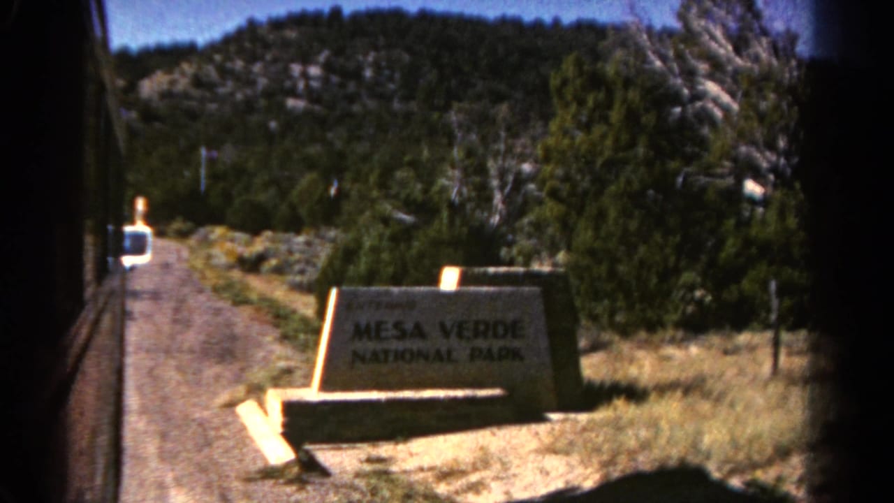 Mesa Verde National Park entrance sign beside a gravel road and evergreen trees.