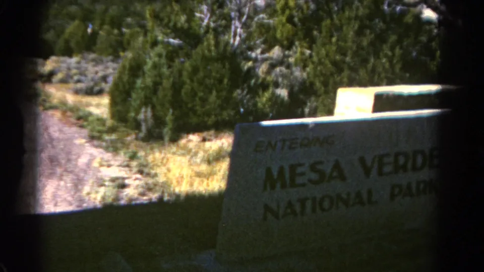 Close 1967 8mm passing view of the Mesa Verde National Park entrance sign in a wooded Colorado roadside setting.