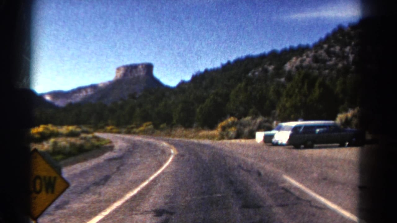 Curving road with a yellow slow sign, parked cars, trees, hills, and a distant butte.