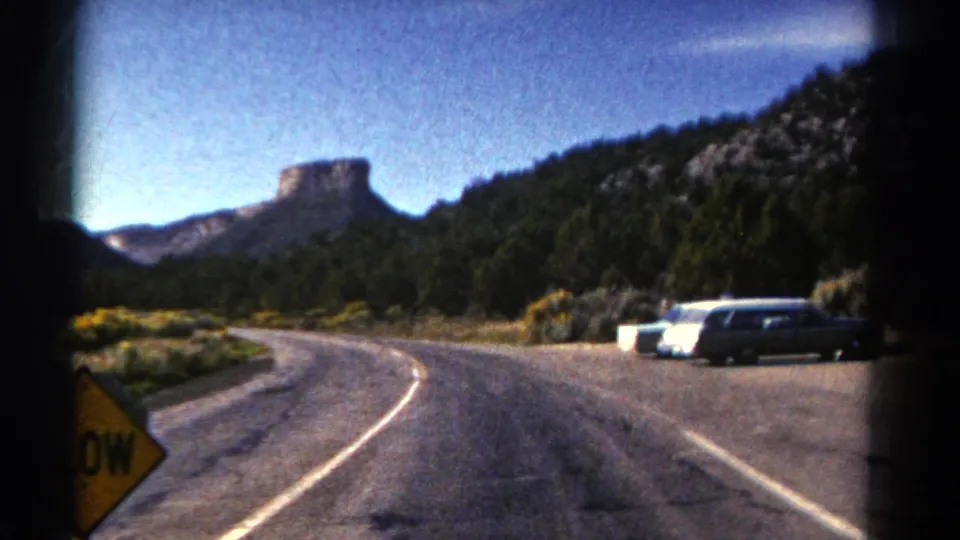 1967 Colorado 8mm frame of a road bending through desert vegetation toward parked cars, wooded hills, and a distant butte.