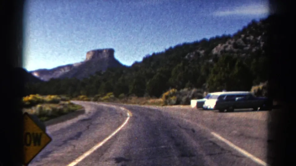 1967 Colorado road-trip frame showing a curving paved road, parked station wagons, dry shrubs, evergreen hills, and a distant butte.