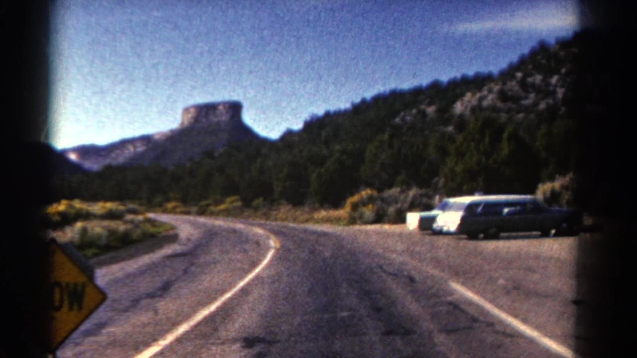 Curving roadside in mountain terrain with parked cars and a partial yellow sign.
