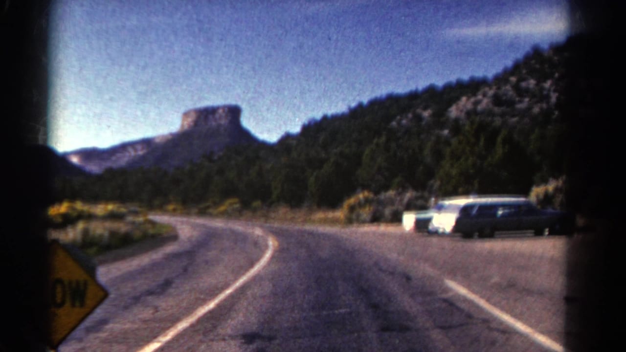 Mountain road curves past a pullout with parked cars and forested hills behind it.