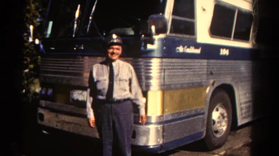 A uniformed adult man stands in sunlight beside a large ribbed metal motorcoach marked with the number 104.