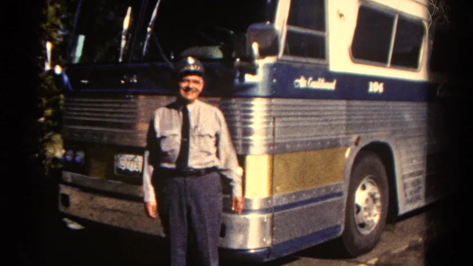 Uniformed bus driver smiles beside a silver and blue coach, with soft 8mm color and dark frame edges.