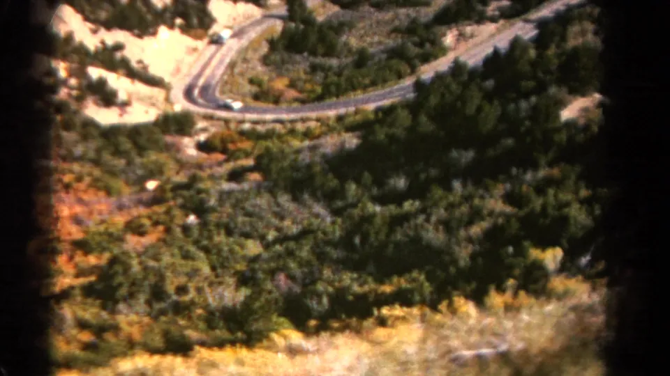 A winding mountain road curves through trees and dry slopes, suggesting the bus journey through Colorado terrain.