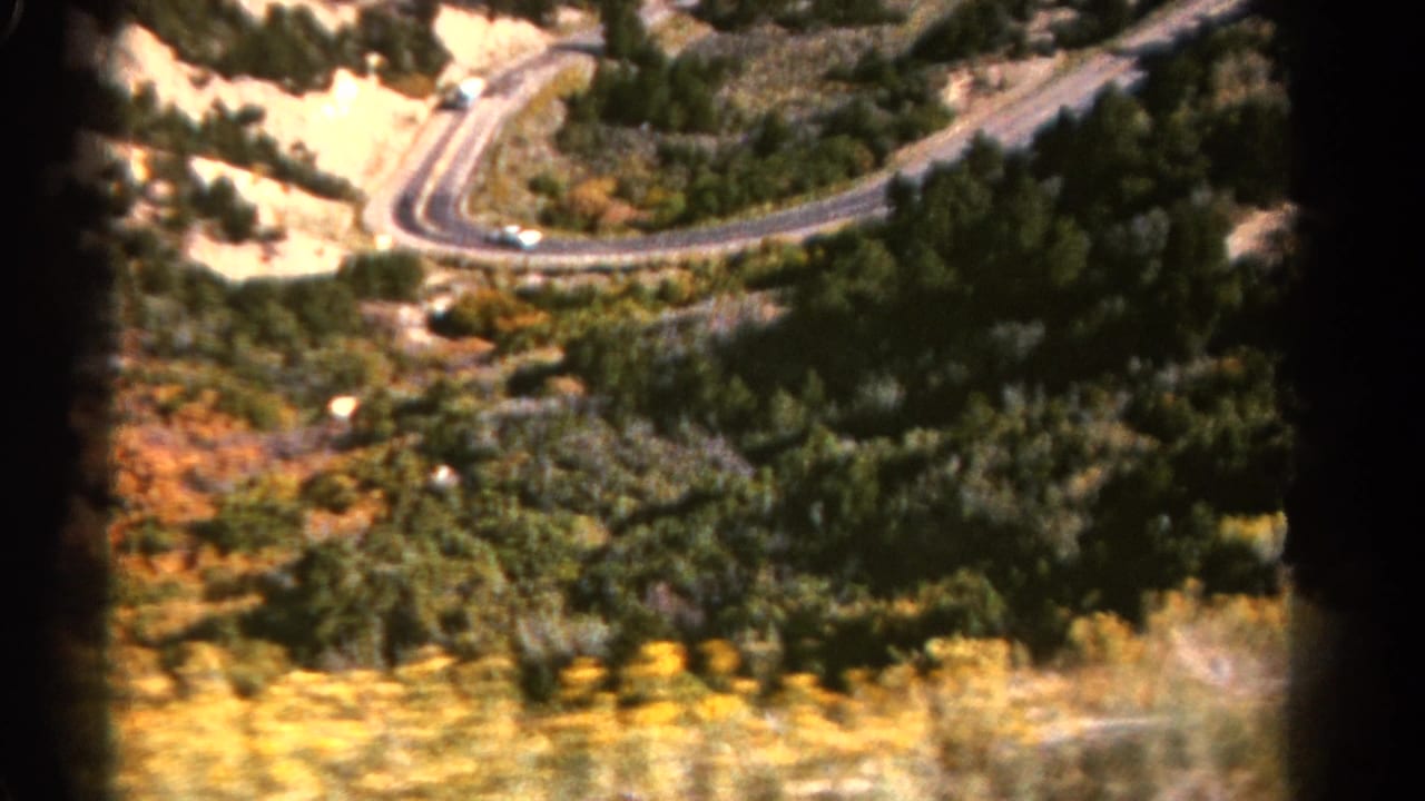 Winding mountain road seen from above through forested hills and dry brush.