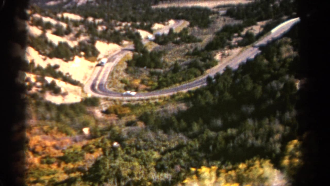 Curving road with small cars passing through wooded mountain terrain.