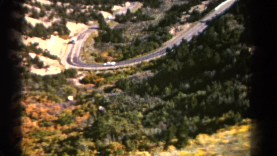 1967 Colorado 8mm view of a mountain road bend bordered by dense green trees, dry vegetation, and bright exposed earth.