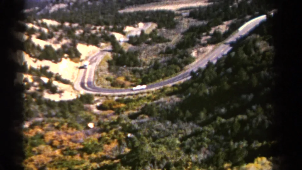 1967 Colorado aerial home-movie frame of cars on a winding mountain road surrounded by forest, scrub, and pale hillside cuts.