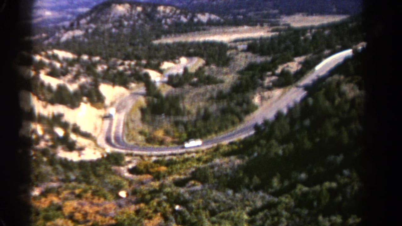 1967 Colorado 8mm frame showing a white bus climbing a curved paved road through pine and rocky mountain terrain.