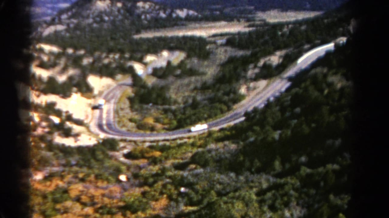 Vintage Colorado home-movie frame of a white bus moving around a broad curve on a mountain road among trees and rocky ground.