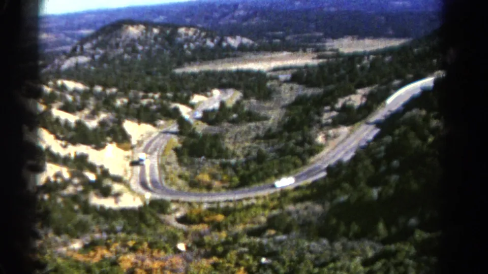 1967 Colorado 8mm still with a white bus on a winding road seen from above, surrounded by trees, scrub, and rocky slopes.