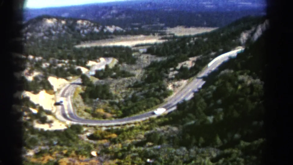 Home-movie frame from Colorado in 1967 showing a white bus on a steep mountain road bordered by forest, scrub, and rocky slopes.