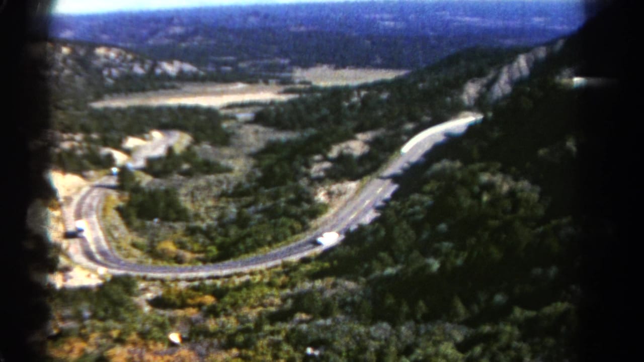 High-angle 1967 Colorado road view with a paved curve, forested hillsides, rocky outcrops, and a small vehicle in motion.