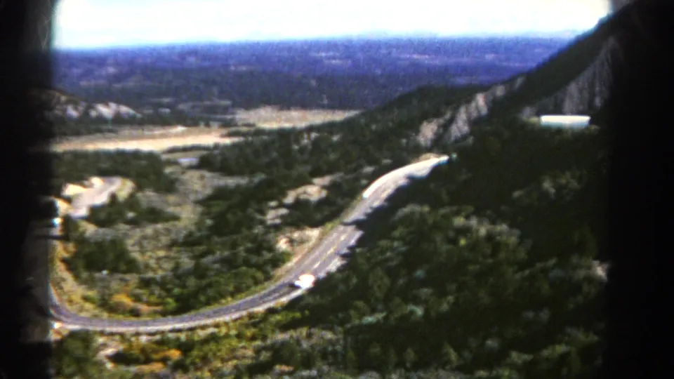 Soft 8mm frame of a Colorado mountain highway descending through trees, with a distant valley and dark film-frame edges.