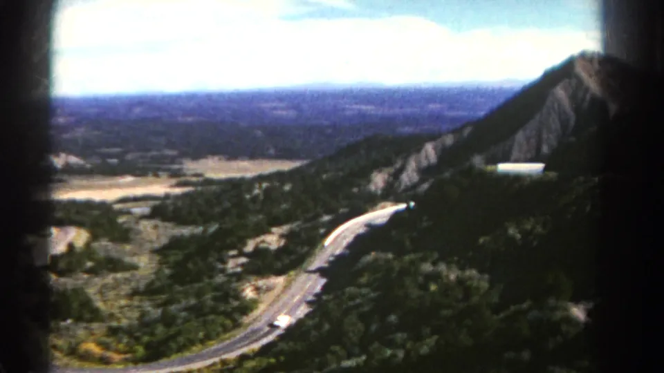 1967 Colorado home-movie frame showing a winding mountain road beside a rocky slope, with a broad valley visible in the distance.
