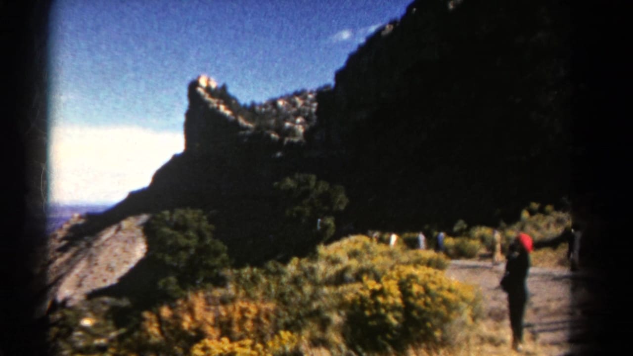 Mountain trail with cliff, blue sky, vegetation, and a visitor in a red hat.