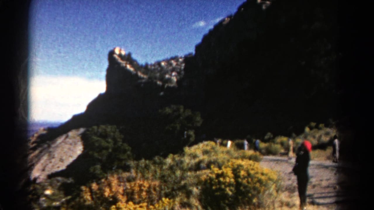 1967 Colorado mountain view with a dark cliff, bright sky, roadside vegetation, and small figures walking near the trail.