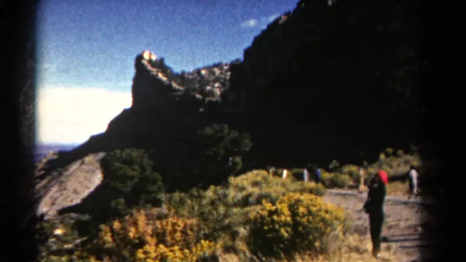 1967 Colorado 8mm frame showing a shadowed cliff, sunlit brush, a curving trail, and visitors spaced along the path.