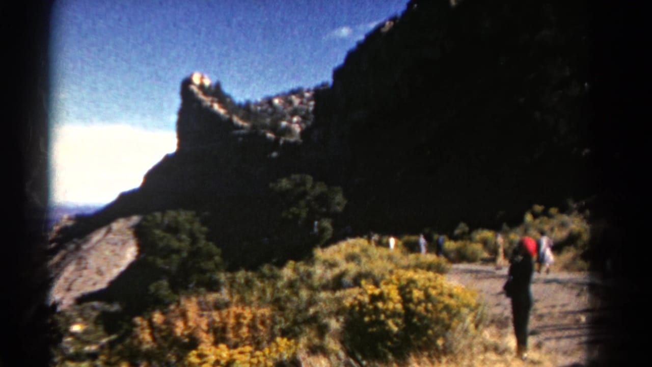 Colorado mountain trail scene with a red-hatted visitor walking beneath a steep rock face in 1967 home-movie footage.
