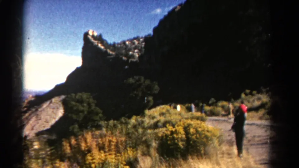 1967 Colorado 8mm footage frame with a visitor on a mountain path, dark cliffs, shrubs, and yellow wildflowers.