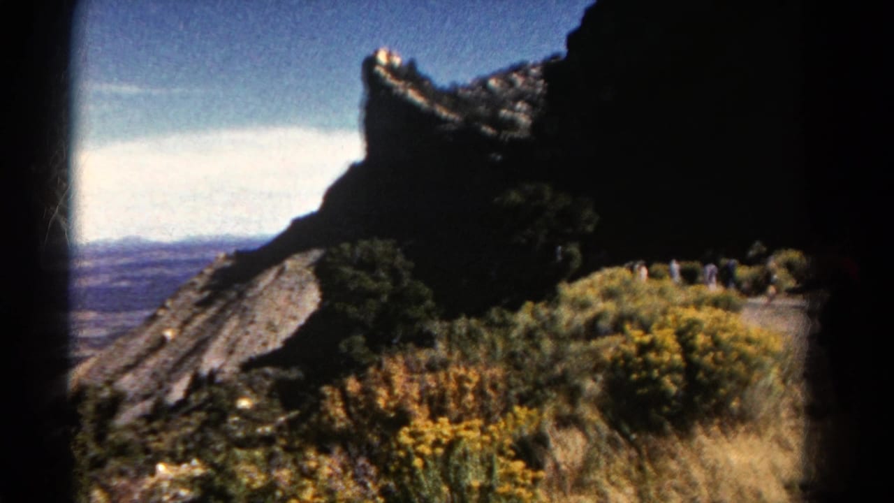 Rocky mountain edge overlooks a broad valley with shrubs and wildflowers in the foreground.