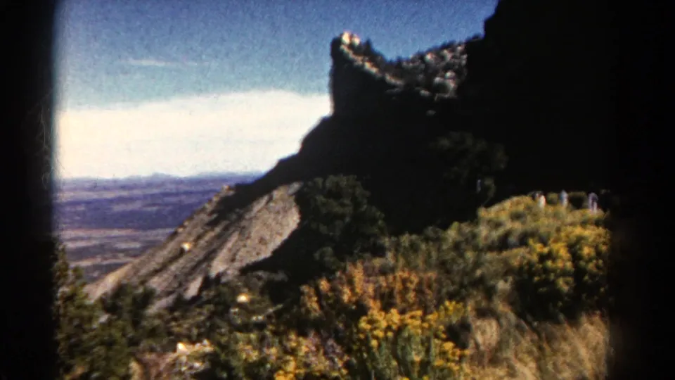 1967 Colorado 8mm frame with a rocky outcrop, hazy valley, foreground brush, and distant sightseers along the path.