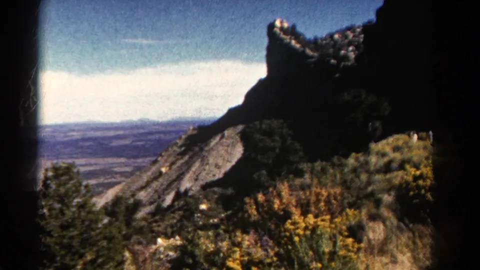 1967 Colorado scenic frame showing a dark rock formation, yellow wildflowers, scrub vegetation, and a distant valley vista.