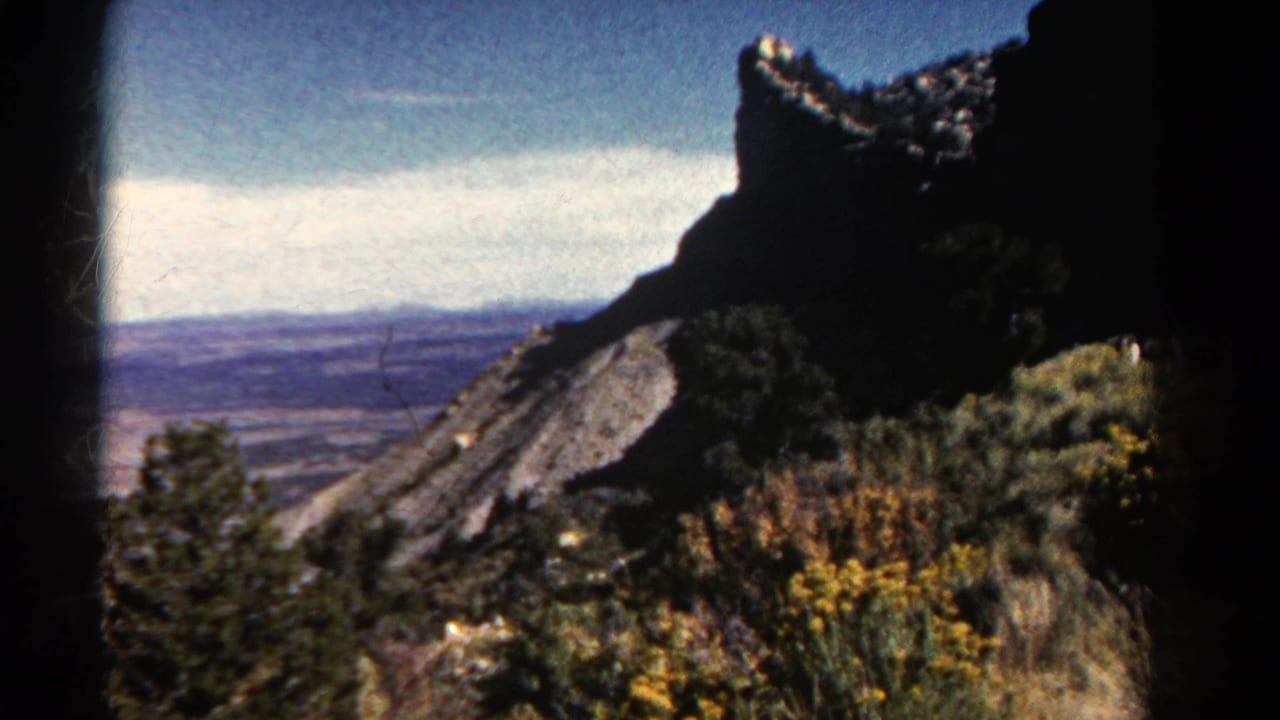 1967 Colorado 8mm frame showing a dark rocky ridge, hillside brush, wildflowers, and a distant valley under a bright sky.