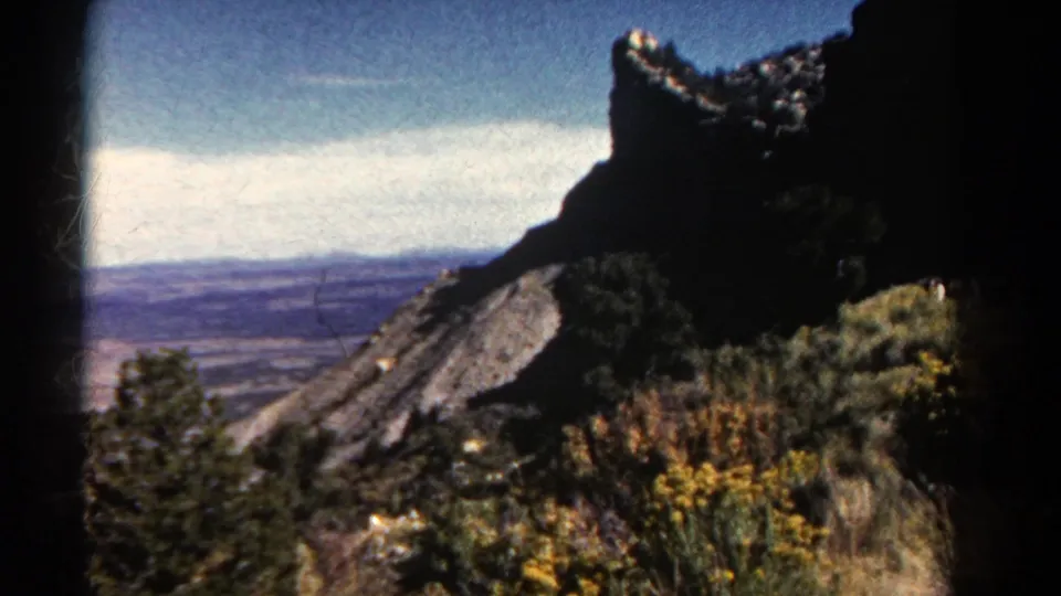 1967 Colorado 8mm frame showing a dark rocky ridge, hillside brush, wildflowers, and a distant valley under a bright sky.