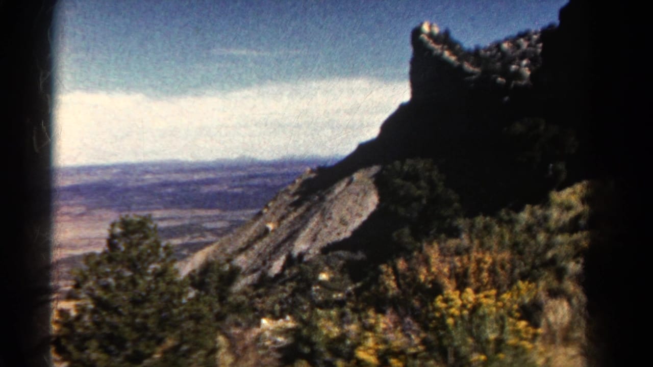 1967 Colorado 8mm frame with a shadowed rock formation, brushy foreground, evergreen tree, and open countryside in the distance.