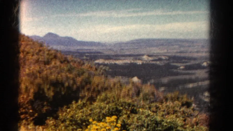 1967 Colorado 8mm frame with foreground shrubs and wildflowers, a wide valley, distant hills, and soft blue sky.