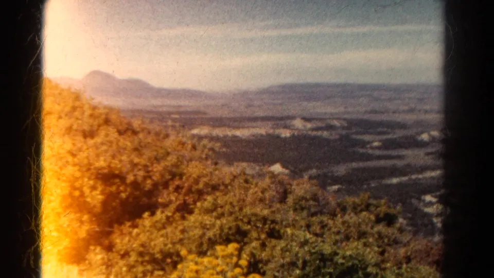 1967 Colorado 8mm frame with warm flare at the left edge, showing hillside vegetation, a broad valley, and hazy mountains.