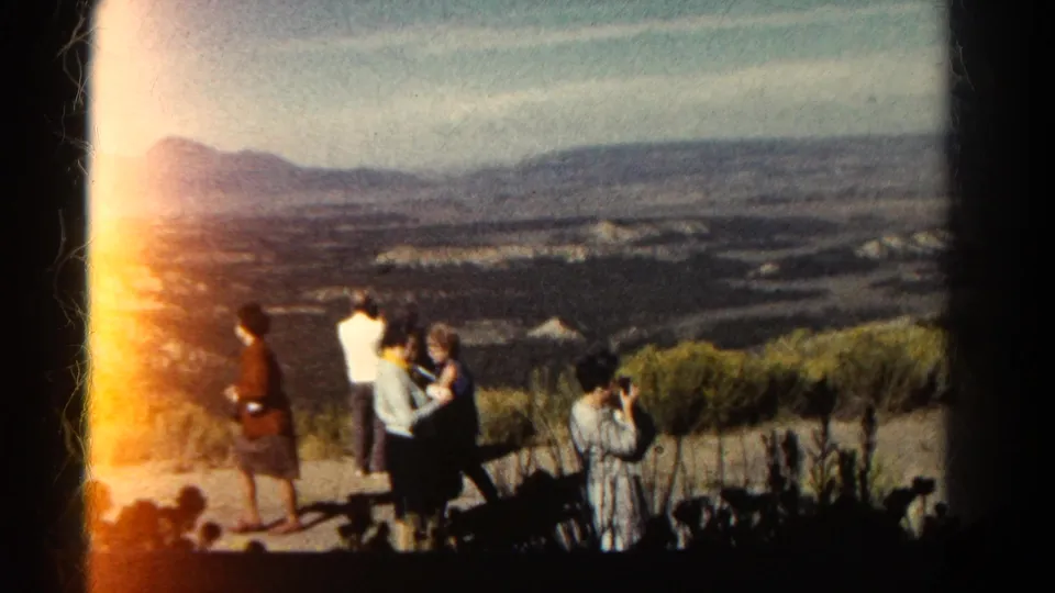 1967 Colorado home-movie frame with adults at a hilltop overlook, one person holding a camera, and distant hills beyond.