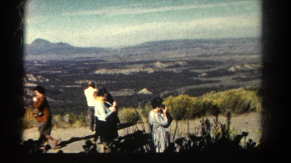 1967 Colorado overlook scene with adults in period clothing, a person using a camera, scrub brush, valley, and distant hills.